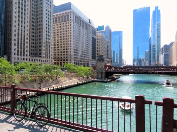 A tour bicycle leaning on the red railing of a bridge with the greenish blue Chicago River below and River Walk seating to the left.