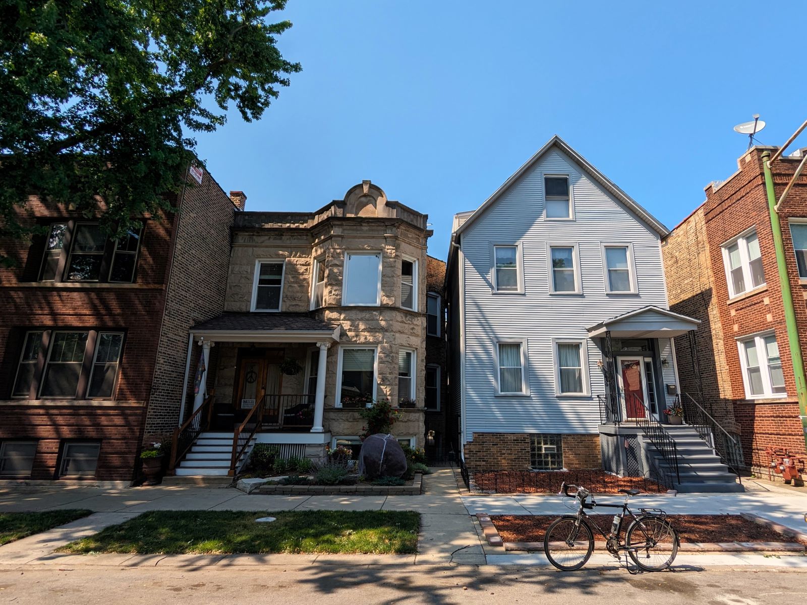 On a neighborhood street a tour bicycle is standing at front of an early 1900s two and half story blue sided wood frame single family home next to a greystone two flat.