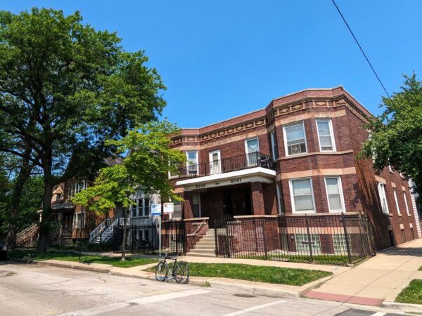 On a tree lined neighborhood street a tour bicycle is standing at front of an early 1900s two story brown brick two flat duplex.