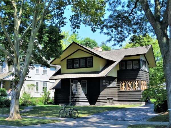 A tour bicycle standing at front of a two story Prairie School home with yellow stucco and second floor horizontal brown wood slats on the first among trees.