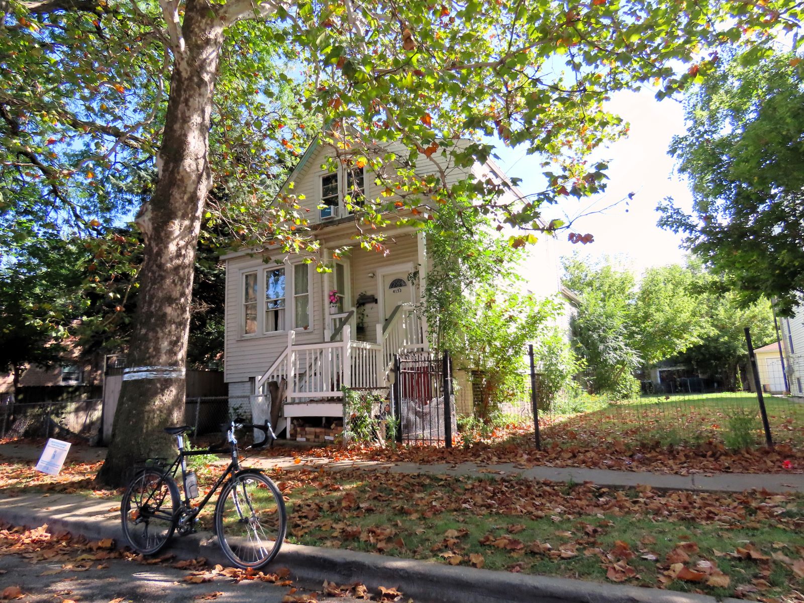 A tour bicycle standing at front of a white wood one and half story worker's cottage under tree coverage and an open side lot.