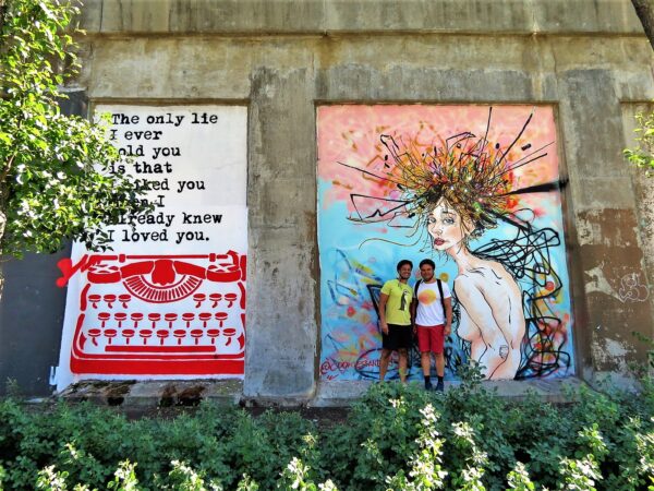 Two CBA bike tour riders posing in front of two murals, the left a stencil of a red typewriter with inspirational text, the right a topless woman with hair of electricity.