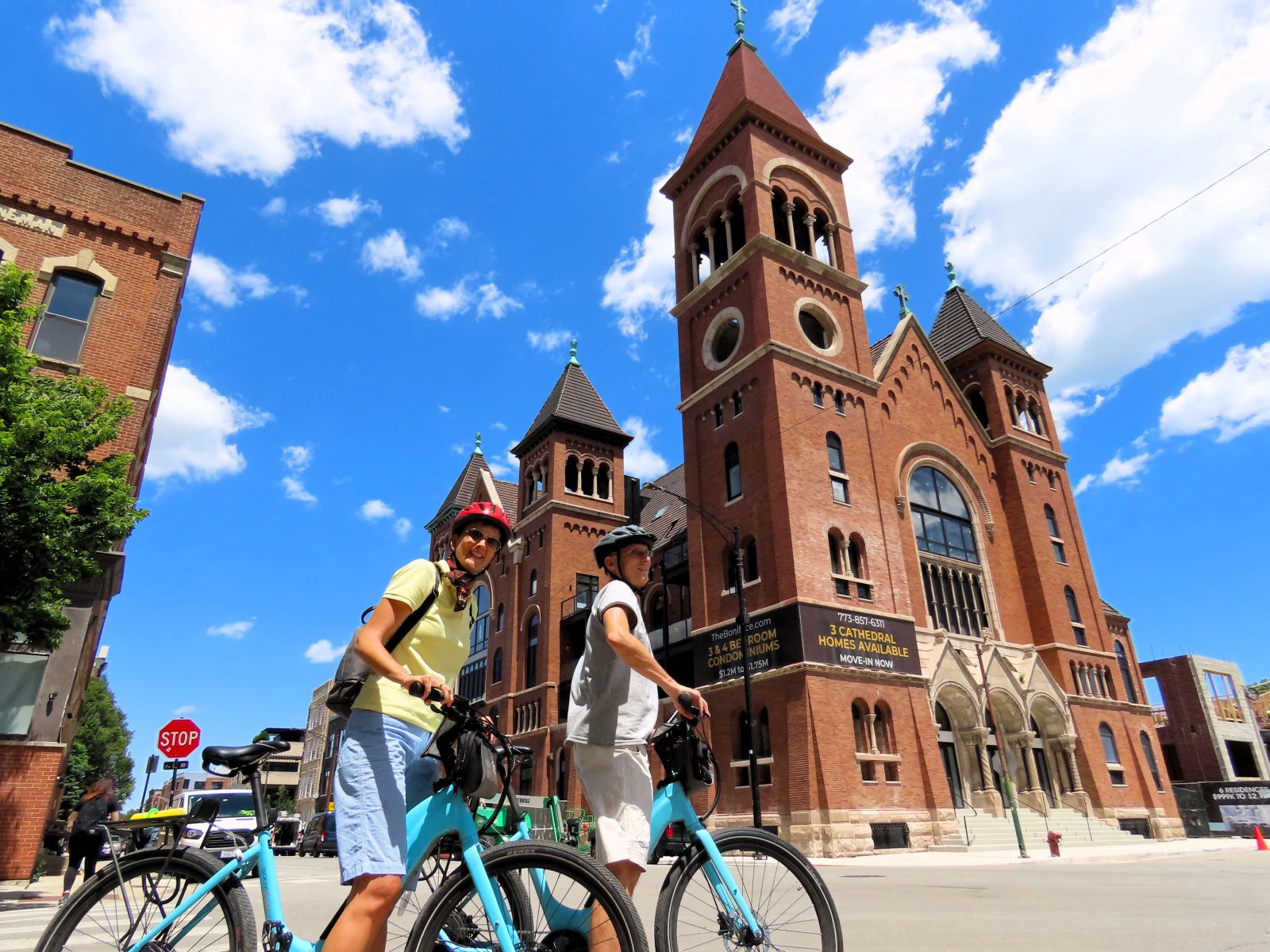 Two CBA bike tour riders posing in front of a large brown brick early 20th Century church being converted in to condos.