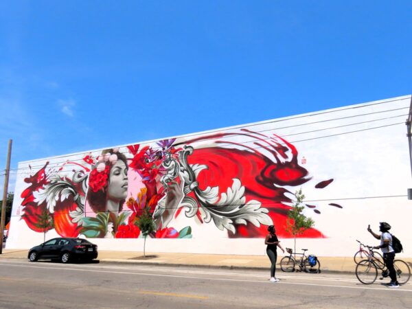 Two CBA bike tour riders posing in front of a large horizontal mural in red and black of a woman surrounded flowers and feathers on a white wall.