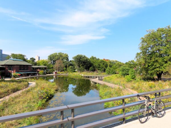 A tour bike leaning on a bridge railing above a pond with a surrounding boardwalk, Prairie School style building, and lush trees behind.