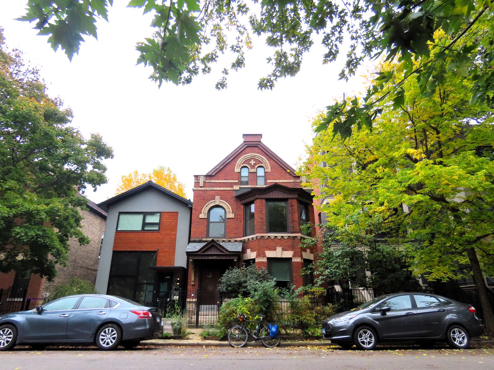 A tour bike between cars in front of an early 1900s reb brick two story home with tan brick decorative design.