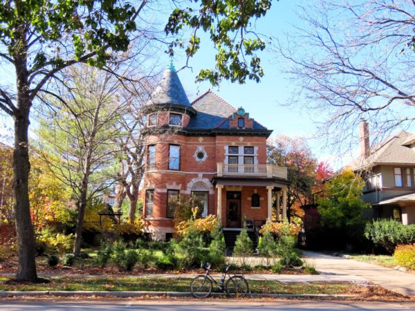 A tour bike standing at front of a two and half story turn of the century brown brick home with a curved wood front porch and turret opposite.