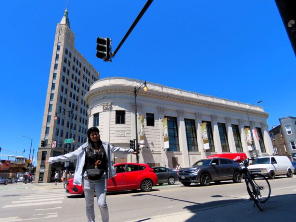 A CBA bike tour rider smiling at the camera with arms wide standing in front of nine story Art Deco corner office building and a two story white terra cotta former bank.