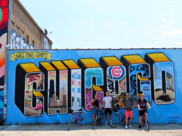 Four CBA riders standing in front of a vintage style Greetings from Chicago postcard mural, smiling at the camera.