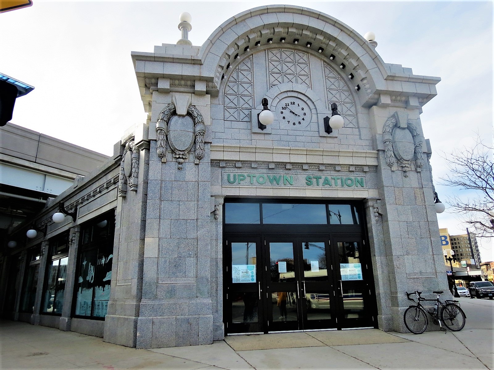A tour bike leaning at the side of the entrance of a grey terra cotta one story corner Beaux Arts train station.