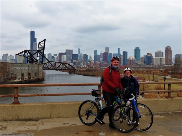 Two CBS tour riders smiling at the camera with the Chicago skyline as seen from the south behind.