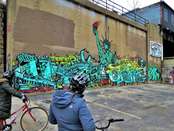 Two CBA bike tour riders looking at an intricate green and yellow Statue of Liberty themed mural within graph.