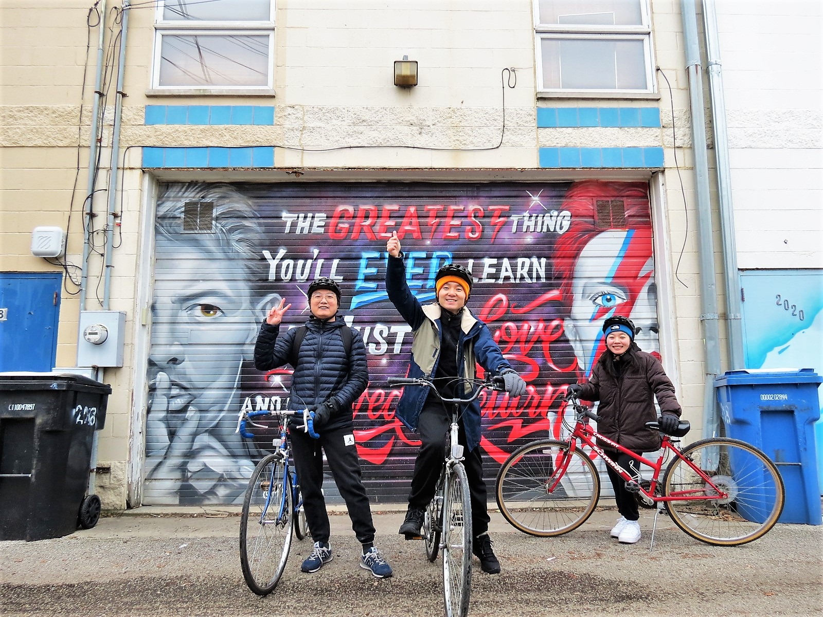 Threee CBA bike tour riders smiling and waving at the camera with a David Bowie garage mural behind.