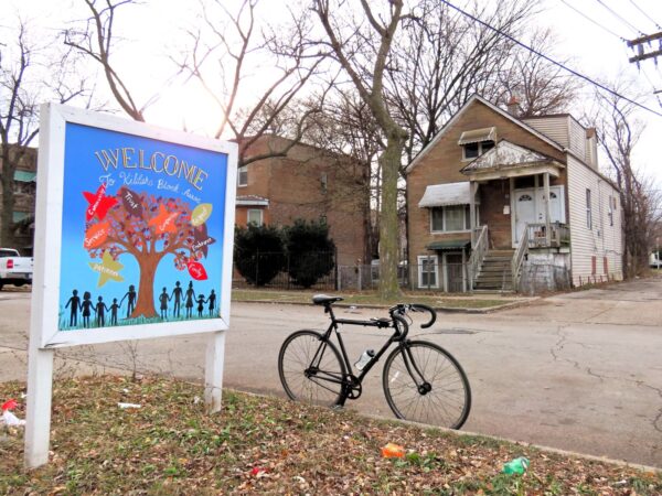 A standing tour bicycle with a colorful wood block sign at left and across the street an aged early 1900s worker's cottage.