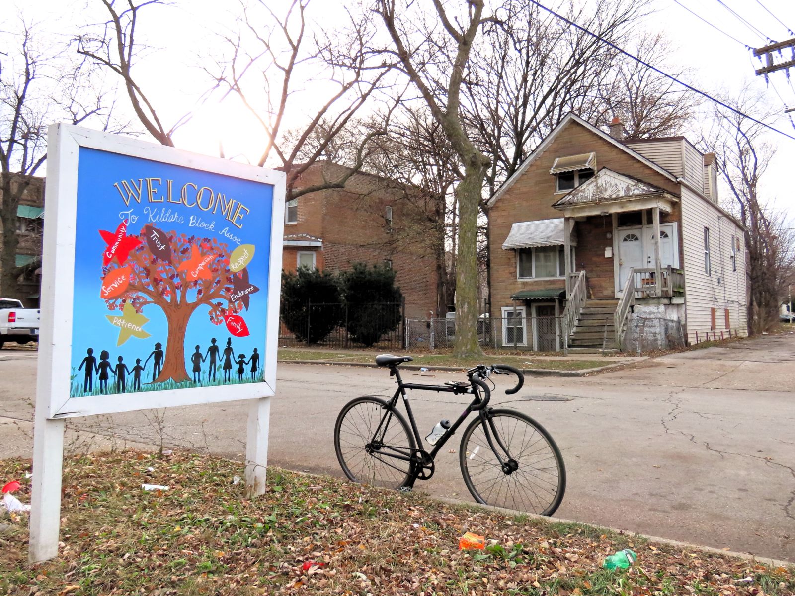 A standing tour bicycle with a colorful wood block sign at left and across the street an aged early 1900s worker's cottage.