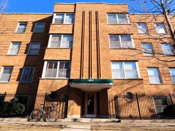 A tour bicycle leaning on a black metal fence around a 1930 four story tan brick Streamline apartments building.