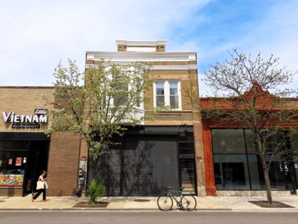 A tour bicycle standing at front of a two story tan brick storefront with a closed first floor roll door and second door bay window and roofline epigram