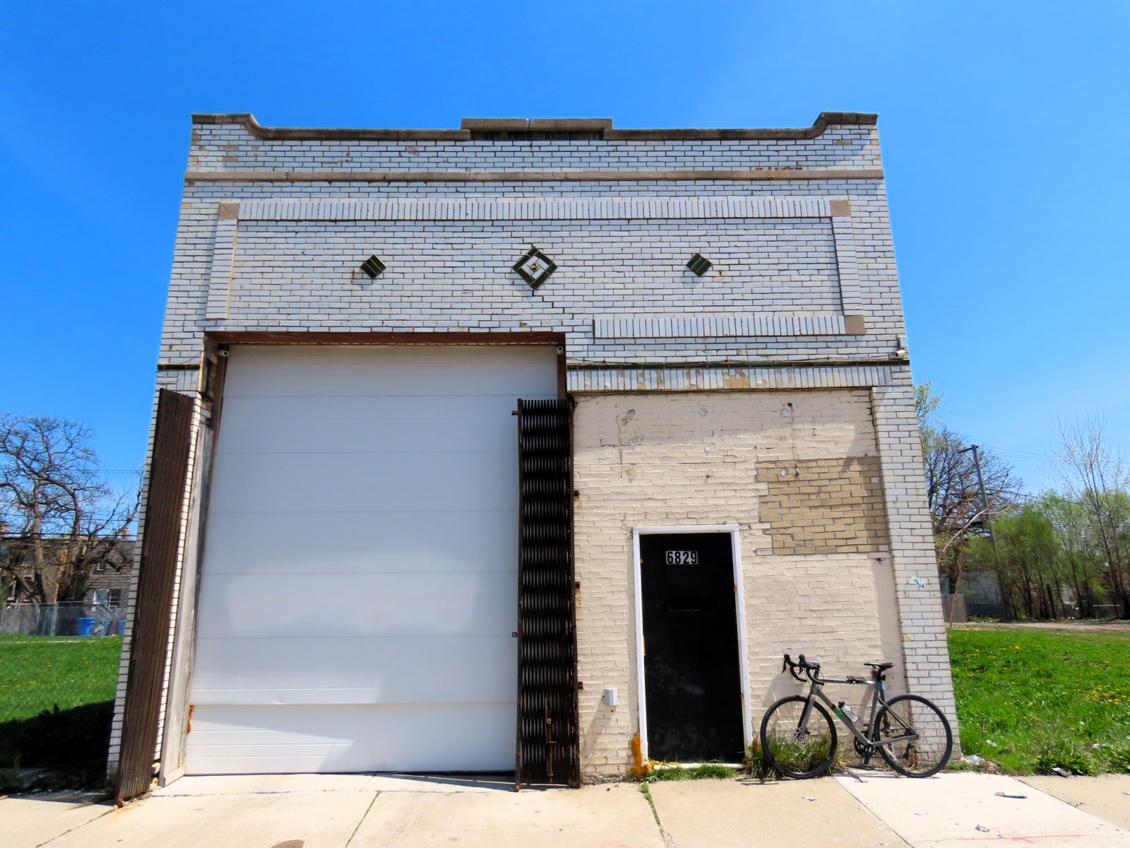 A tour bicycle leaning on the front of one story green and white glazed brick garage with a roll bay door on the left.