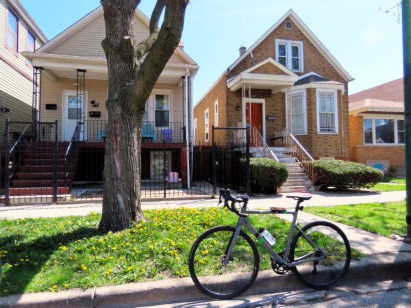 A tour bicycle standing at front of two early 1900s one and half story worker's cottages, the right tan brick, the left wood obscured by a tree trunk.