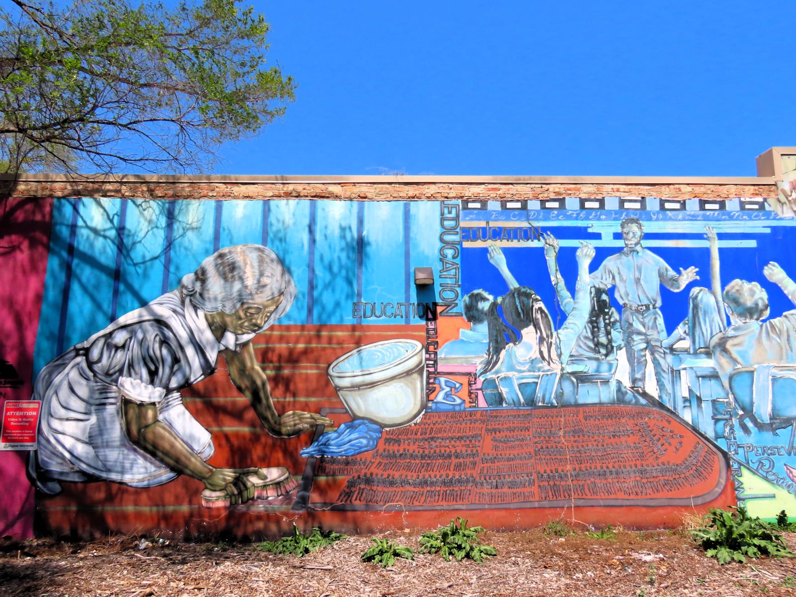 A blue based mural of at left an older black woman on her knees scrubbing clean a board covered y human figures arranged in the same fashion a drawing of 18th century slave ship holds and at right a Black male teacher addressing a class seen from behind with all of their hands raised.