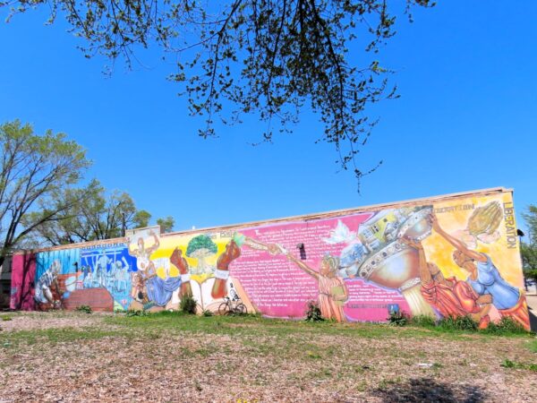 Across an empty lot a tour bike is leaning on a blue, yellow and pink based mural with two chained together black hands and other captive and liberation images.