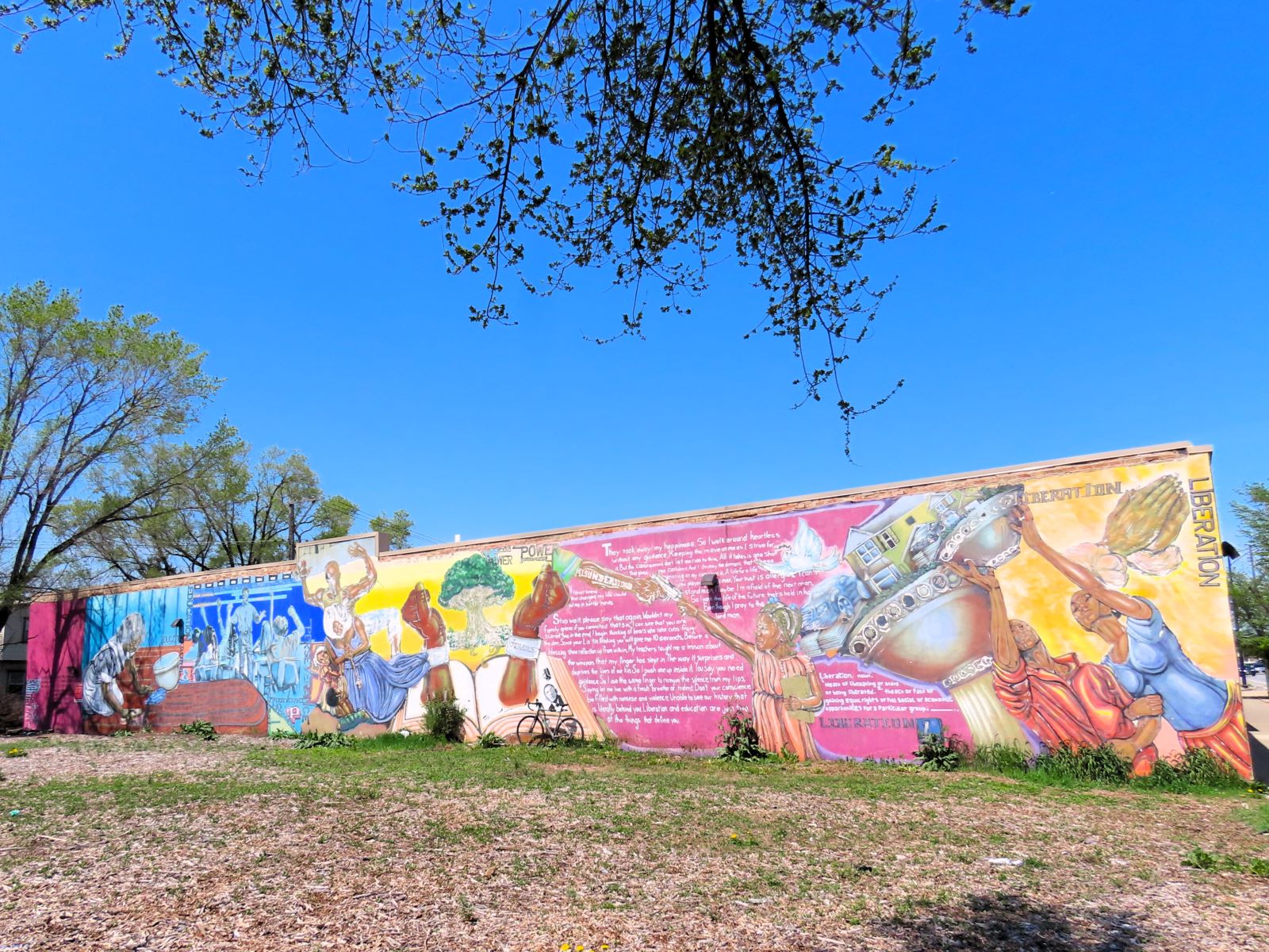 Across an empty lot a tour bike is leaning on a blue, yellow and pink based mural with two chained together black hands and other captive and liberation images.