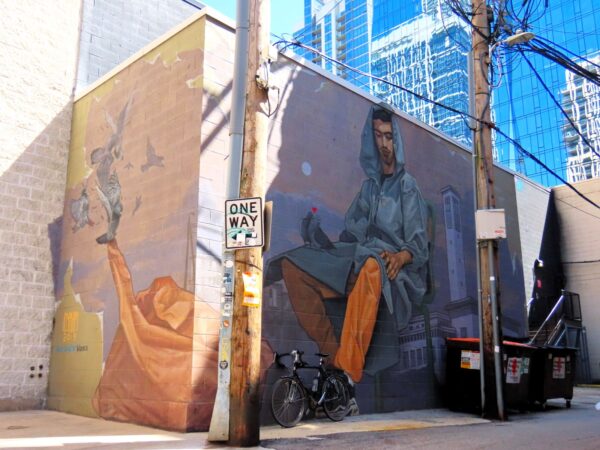 A tour bike leaning on a lavender based mural of a seated man with hood up and tan pants.