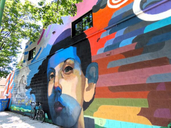 A tour bike leaning on a muraled wall, layers of color at right that lead to a boys face with forehead and chin covered in blue, then a cloudy blue sky left