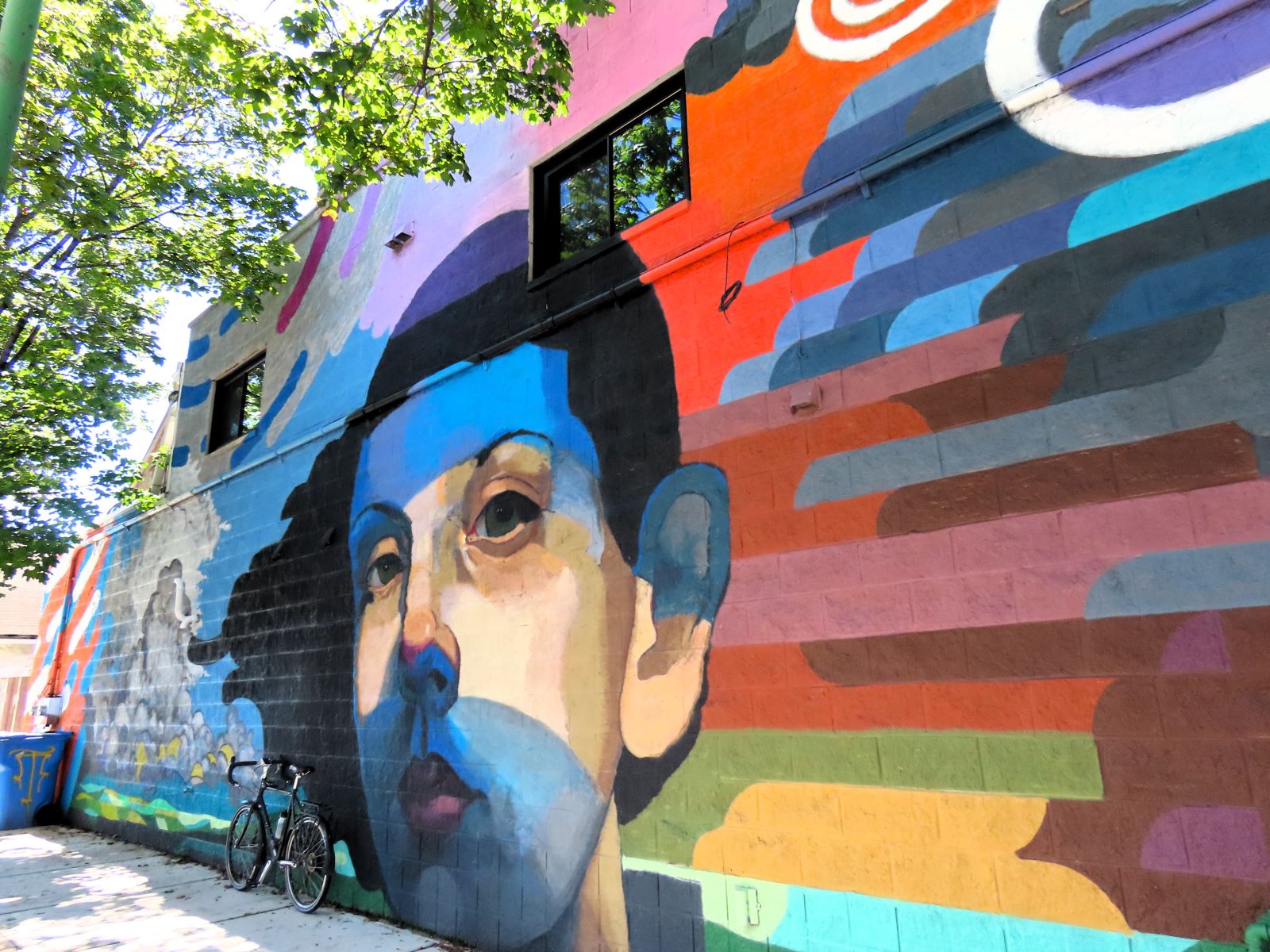 A tour bike leaning on a muraled wall, layers of color at right that lead to a boys face with forehead and chin covered in blue, then a cloudy blue sky left