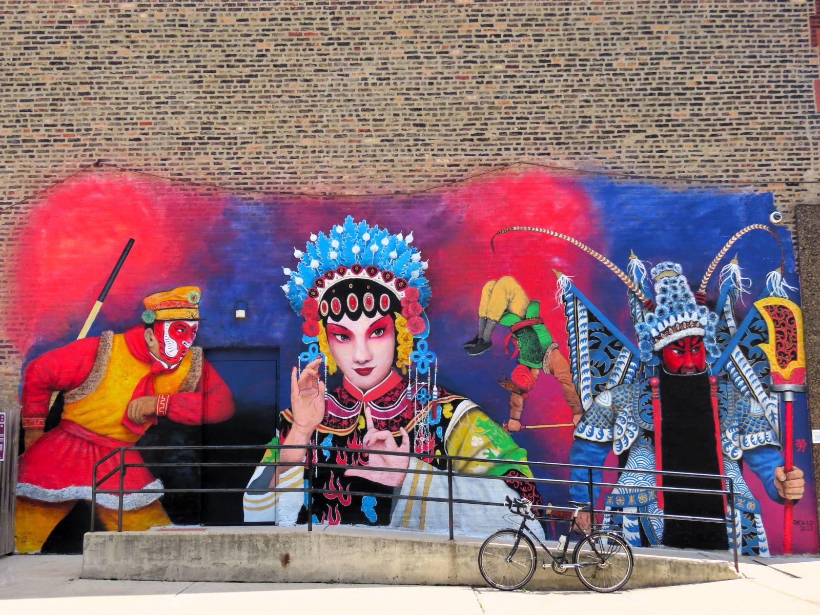 A tour bike leaning on a metal handrail in front of a mural showing four figures from Chinese opera. 