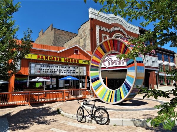 A tour bicycle standing in front of a rainbow wheel sculpture in front of a red brick and white terra cotta theater and a one story orange painted restaurant with thin yellow and red marquee that reads Pancake House.