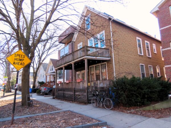 A tour bike on a city neighborhood sidewalk leaning in front of a two and half story early 1900s wood frame two flat with a wide front wood porch on both floors.